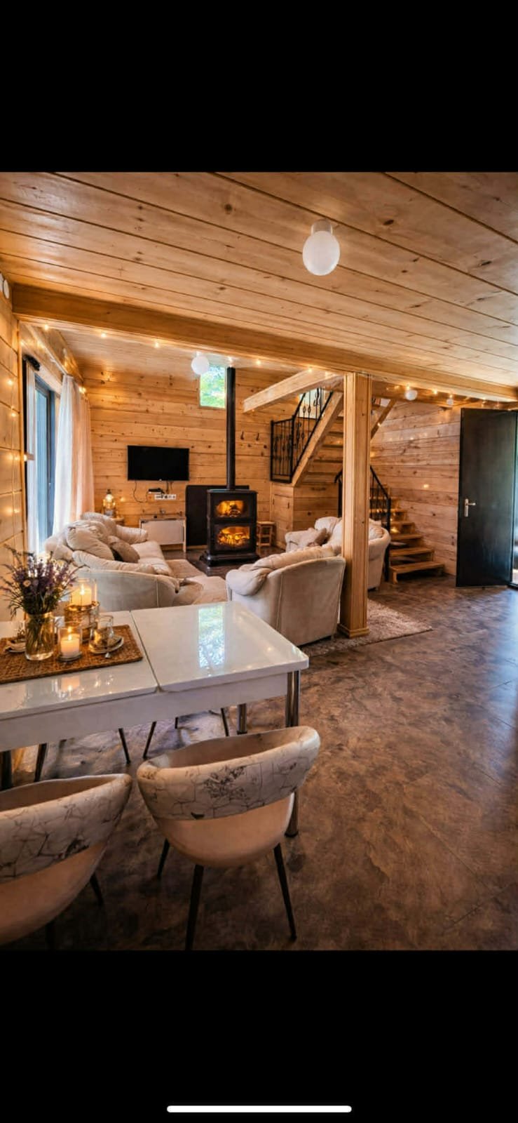 Modern wooden cabin interior with dining area, cream furniture, fireplace with stove, wooden ceiling, and staircase in background