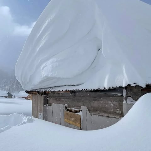 Wooden cabin heavily buried under massive snow drifts during winter storm, with snow piled high above the roof