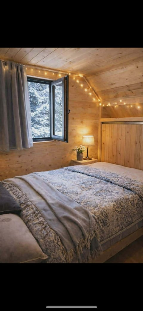 Cozy wooden cabin bedroom with a bed, warm string lights, window overlooking snowy trees, and a bedside lamp