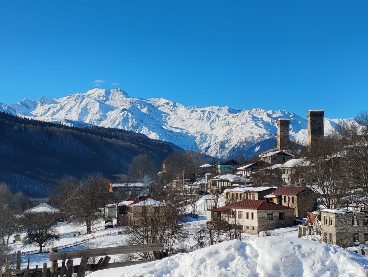 Alpine village with historic stone buildings and church towers set against snow-capped mountains under clear blue sky