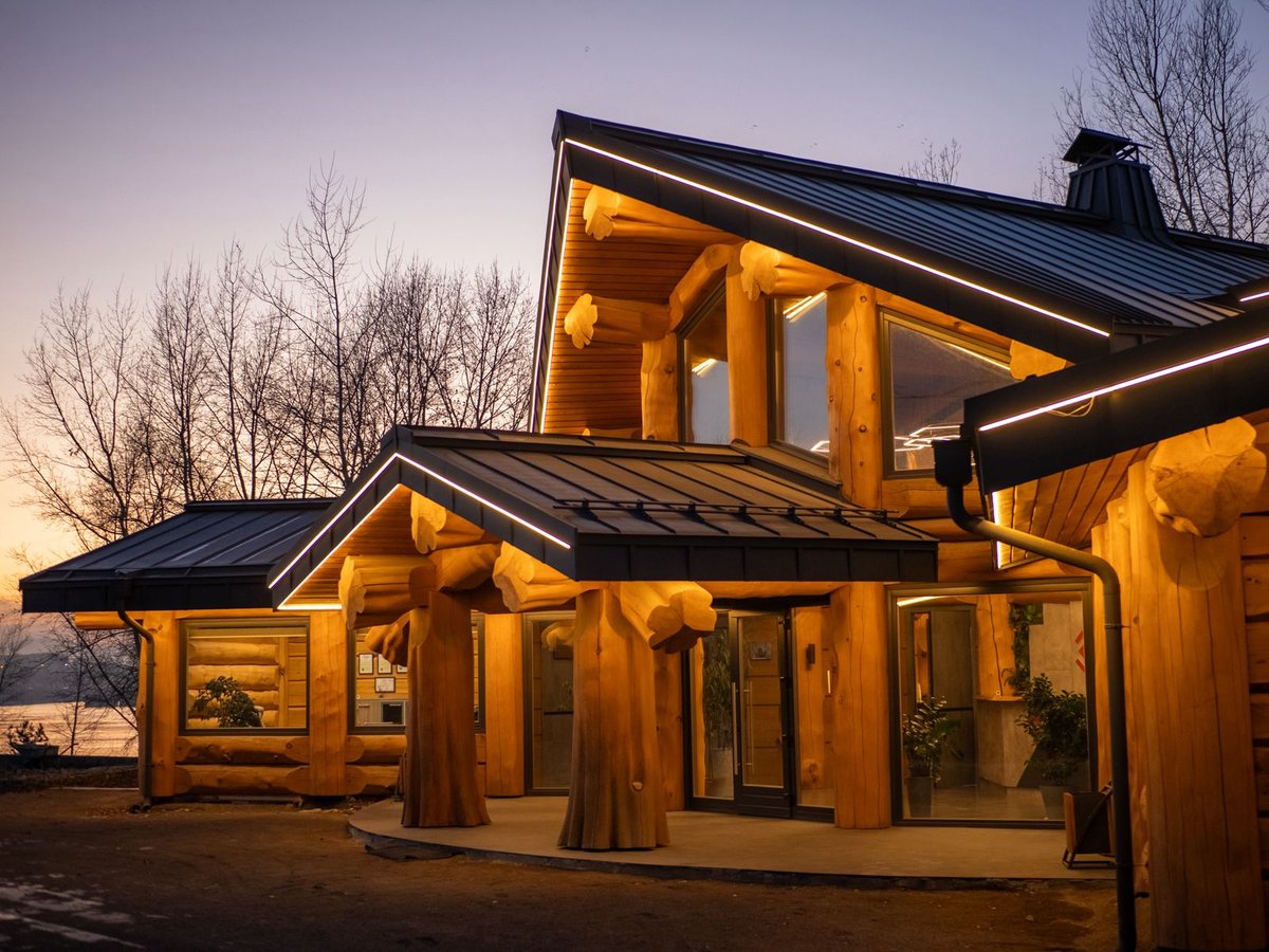 Modern log cabin with illuminated interior at dusk, featuring large windows and metal roof against bare trees